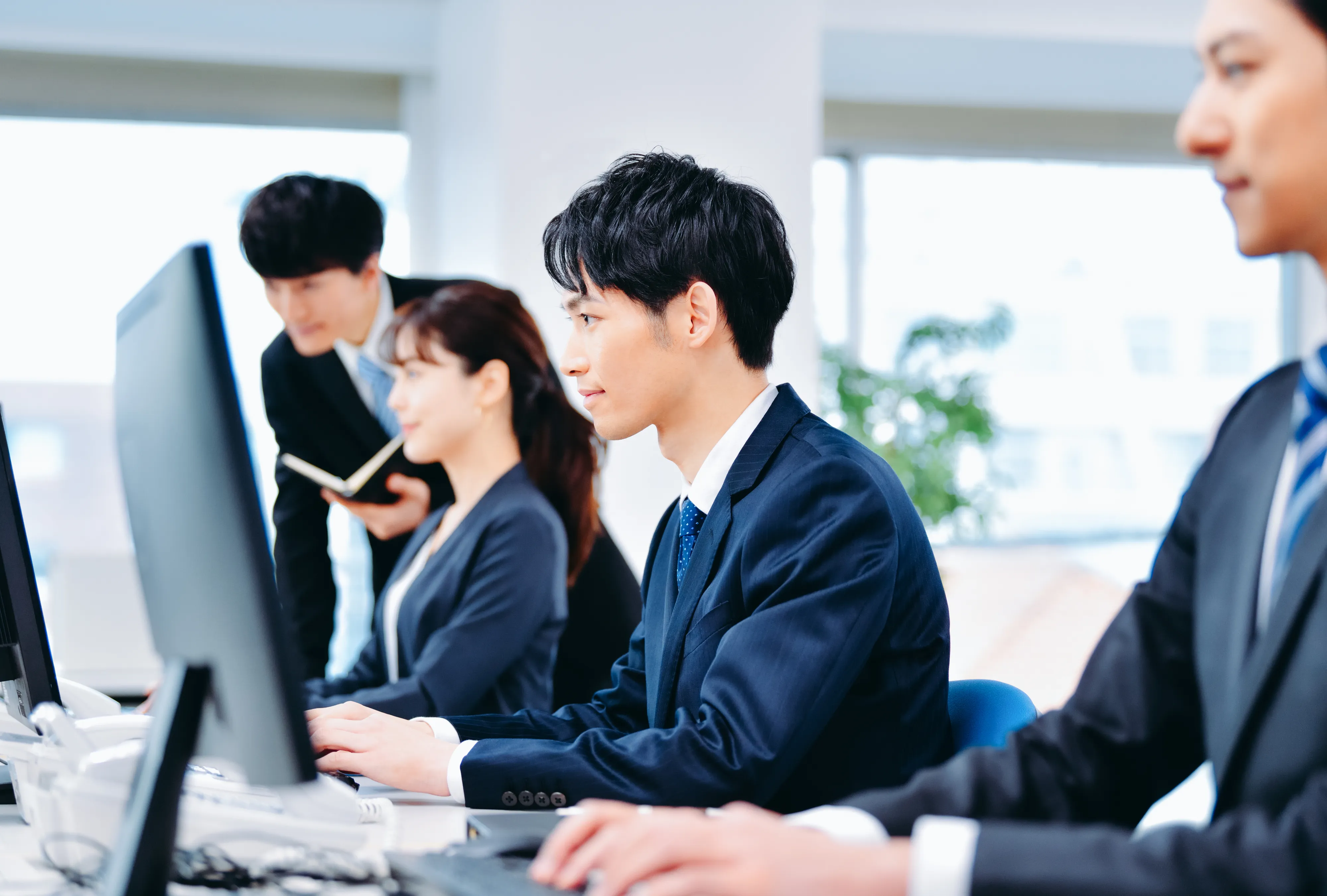スーツ姿の人たちがオフィスでパソコン操作をしている様子